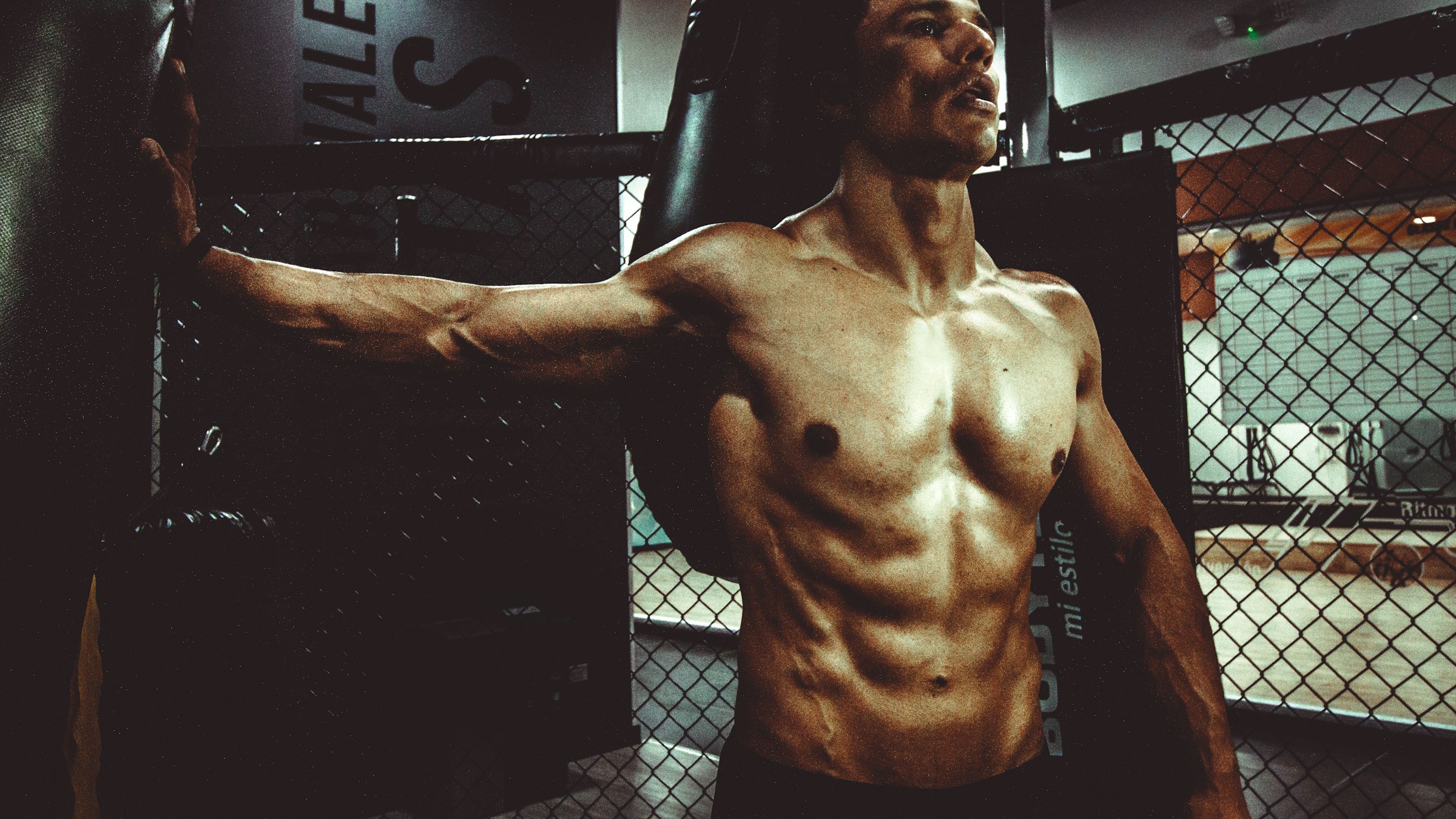 A shirtless muscular man with his right arm out to the side and hand braced against a boxing bag, stretches his pectoral muscle